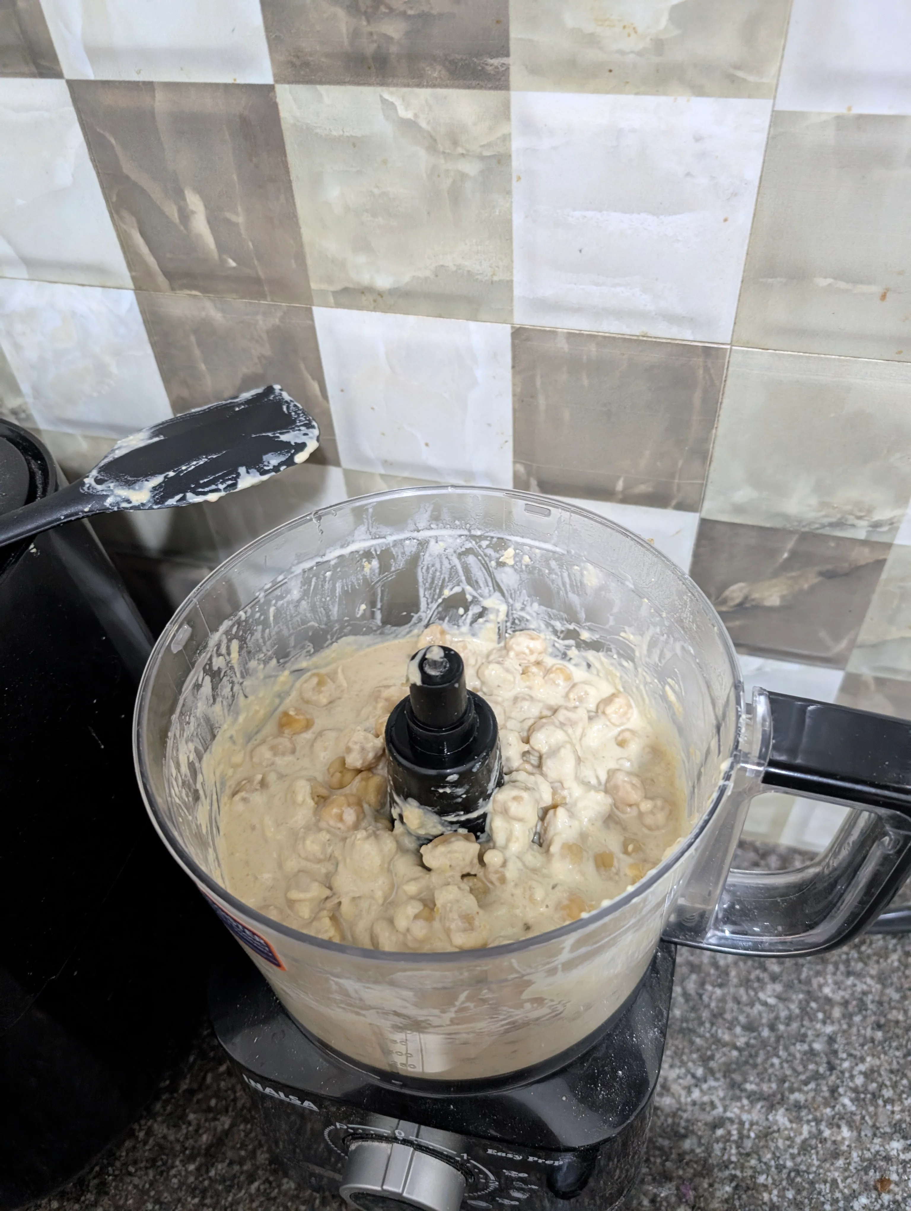 A food processor with blended chickpeas inside it, and a spatula sitting a little to the left that has clearly been inside the food processor. You can still see a bunch of solid chickpeas that are yet to be grounded down, floating in a slightly off white paste of ground chickpeas.