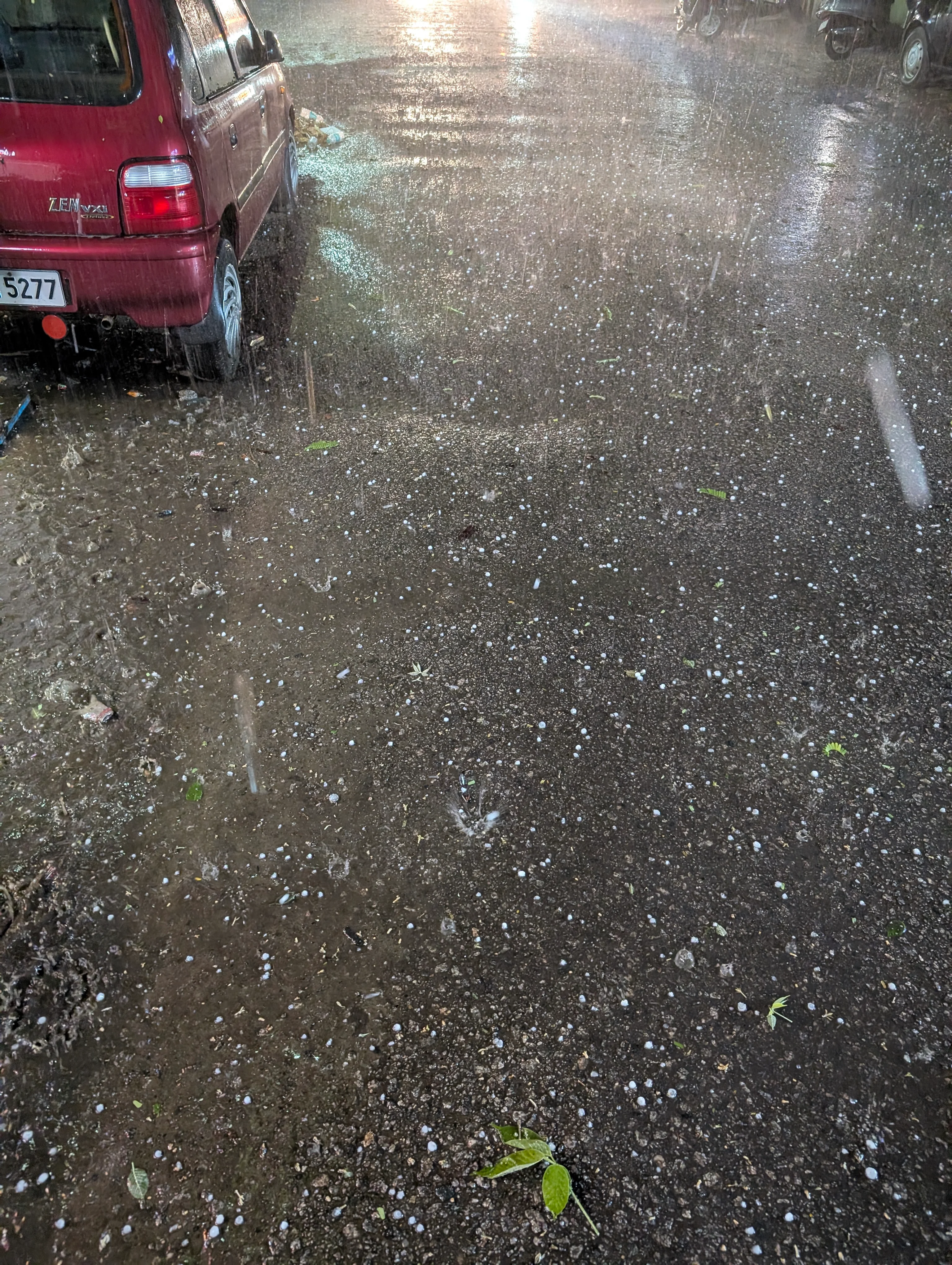 A short video of rain and hail falling on a slightly waterlogged asphalt road. A red Maruti Suzuki Zen car is visible in the top left corner, with a partial number plate.