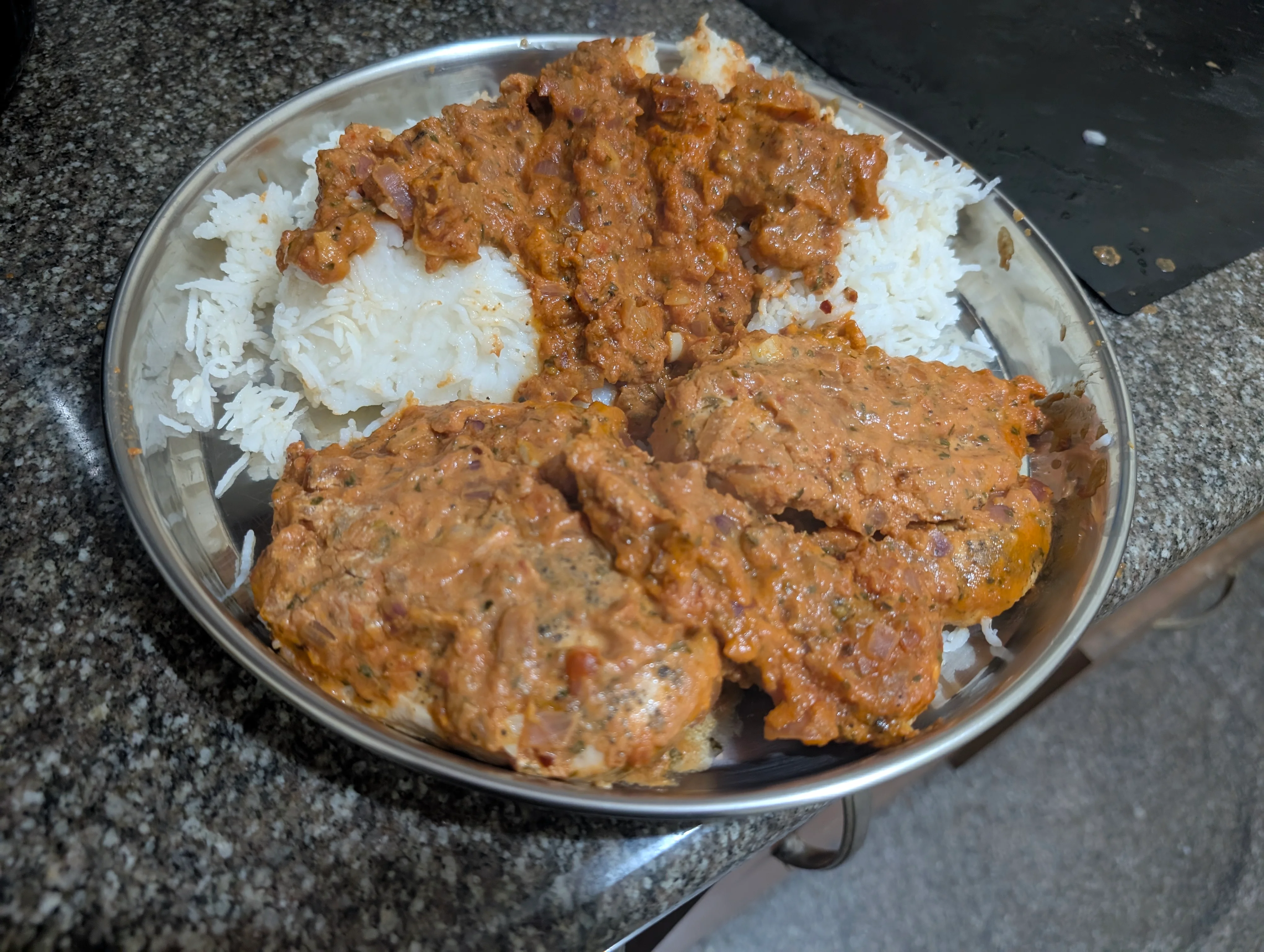 A plate of two chicken breasts and some rice with gravy over it. The gravy is reddish-orange in color due to the tomatoes, turmeric and heavy cream coming together. There are also bits of onion visible in the gravy.