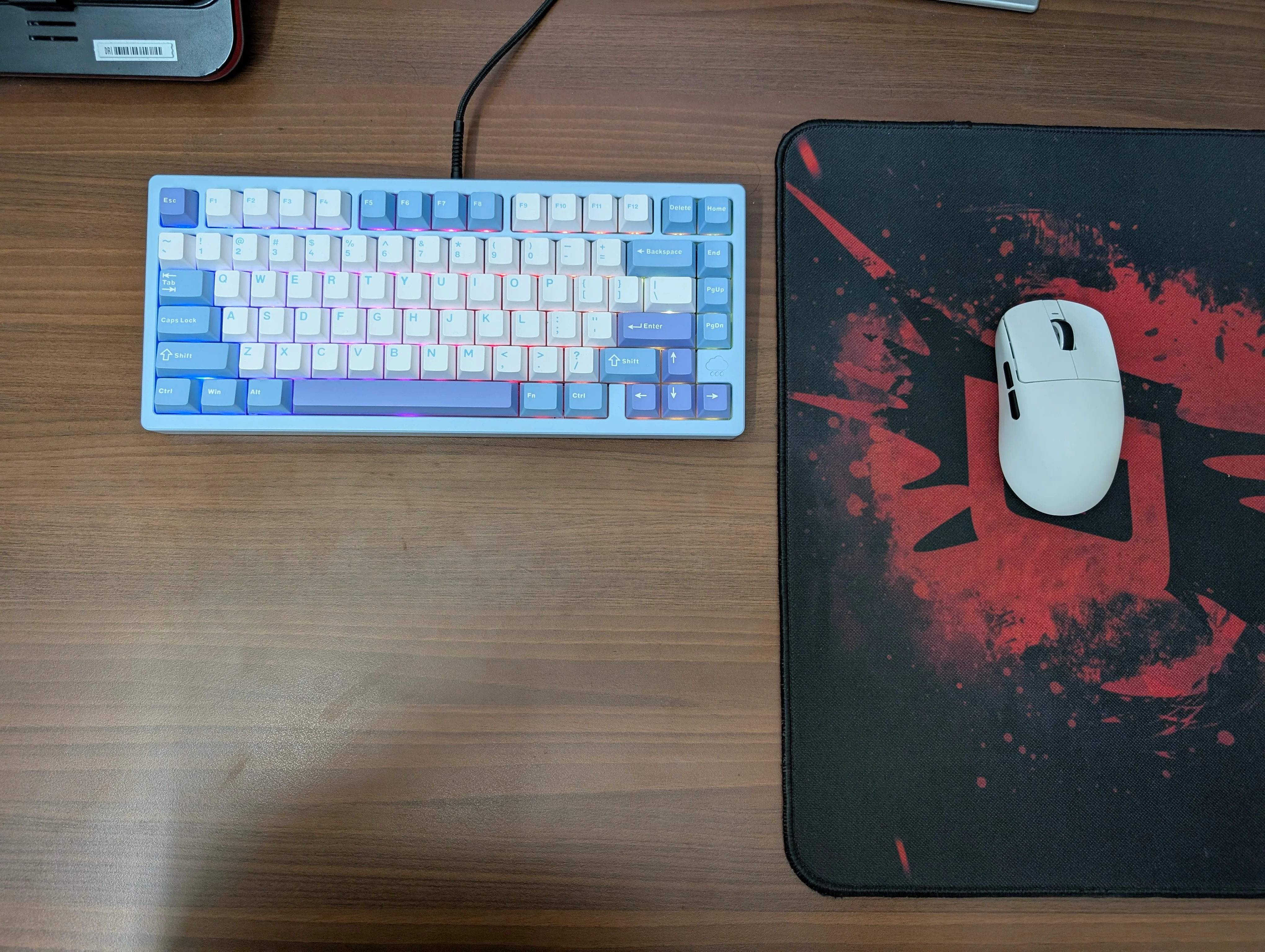 The new keyboard and mouse on top of a wooden particle board desk. The keyboard has an aluminum frame coated in blue, with a mix of blue, purple and white keys with white being the most common one. The mouse rests on a cheap RedGear mousepad.