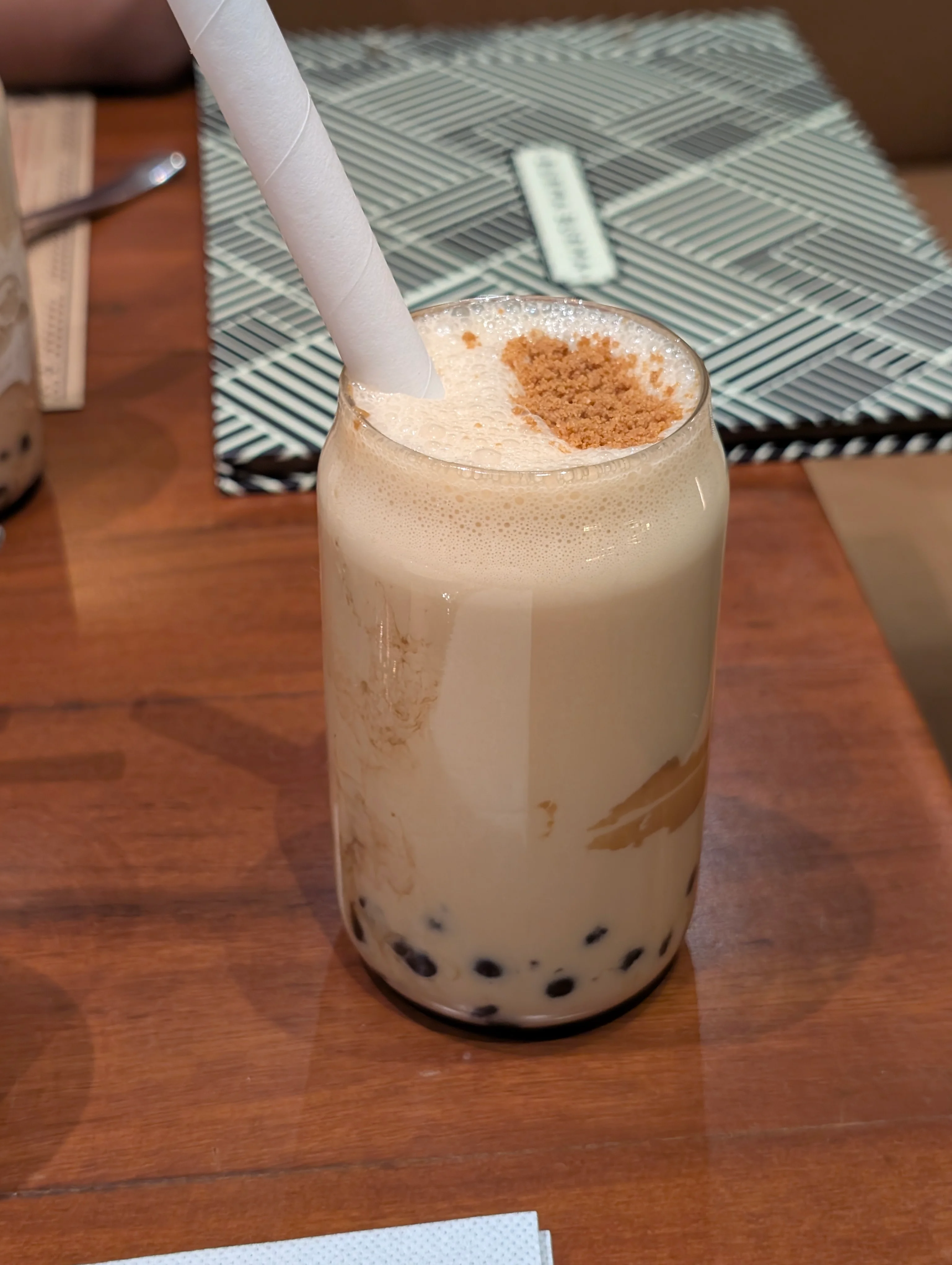 A glass of boba tea with a thick straw in it. The black boba is visible near the bottom, and the drink is a very faded cream color with some biscoff biscuit crumbs on top as a garnish.