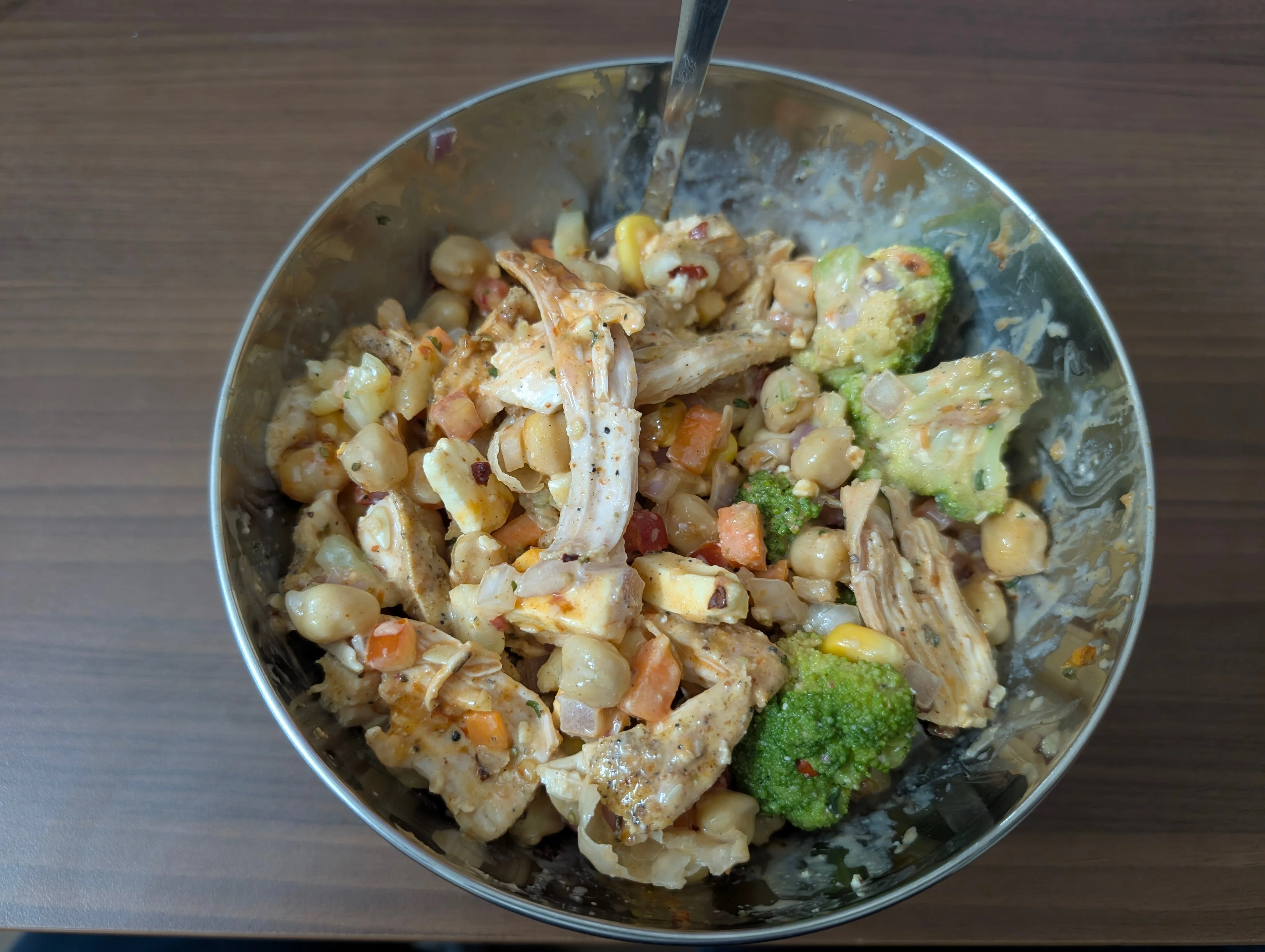 A chickpea salad bowl containing strips of air fried chicken, some impeccably blanched broccoli, cubes of cottage cheese, corn, onions, and tomatoes. All of this is coated in a greek yogurt based sauce which had some random spices added to it.