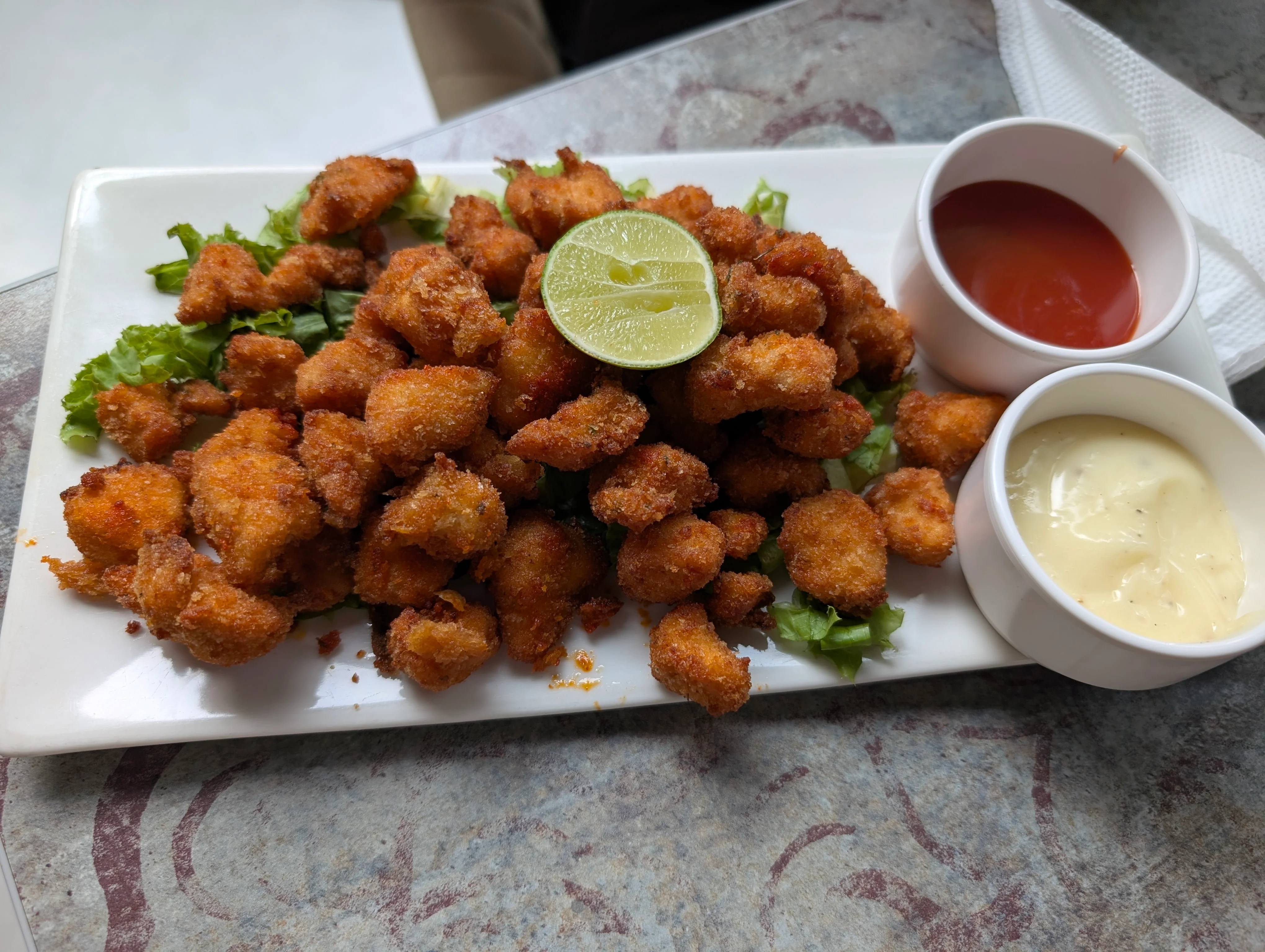 A plate of fried chicken popcorn with small bowls of mayonnaise and ketchup. There's a lemon half sitting on top of the chicken, which itself is on thin bed of lettuce.